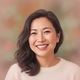 A smiling East Asian woman in her 30s, with a genuine, happy expression, against a soft, warm background.