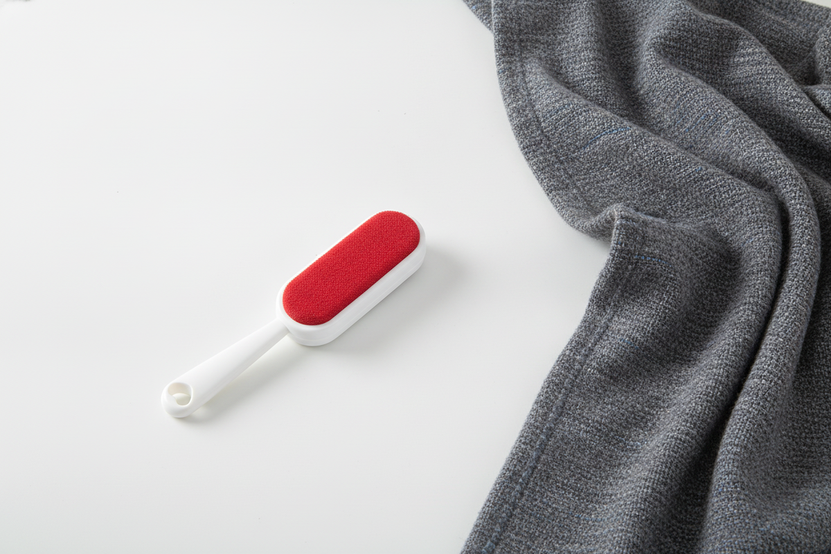 A double-sided, self-cleaning pet hair remover rests on a white surface next to a charcoal throw blanket with slate blue acce